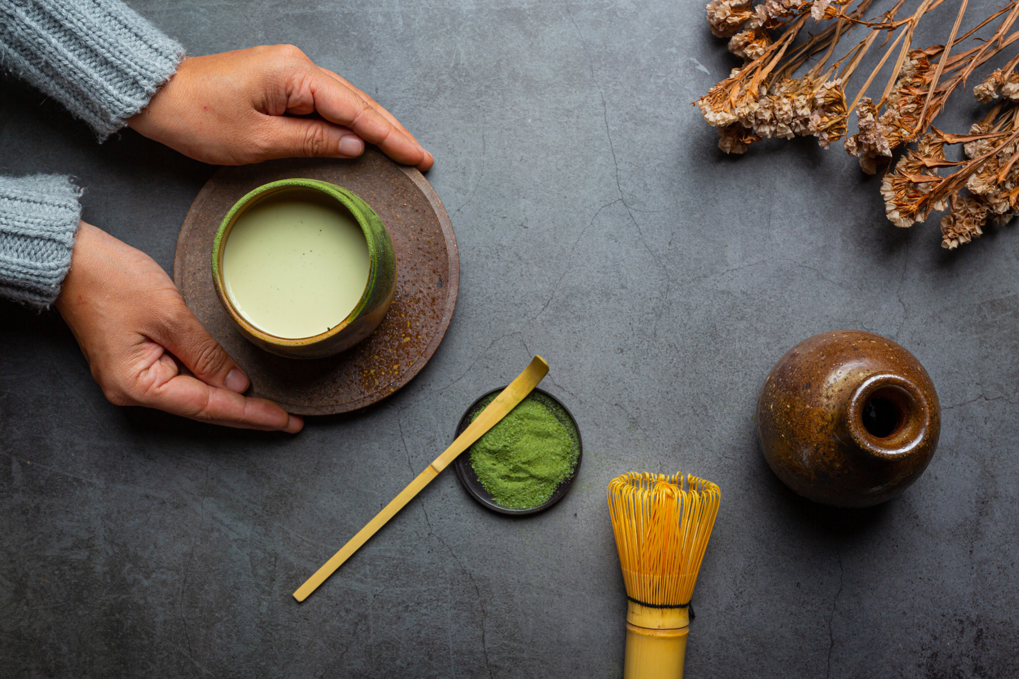 Hands holding a cup of matcha with whisk and green tea powder